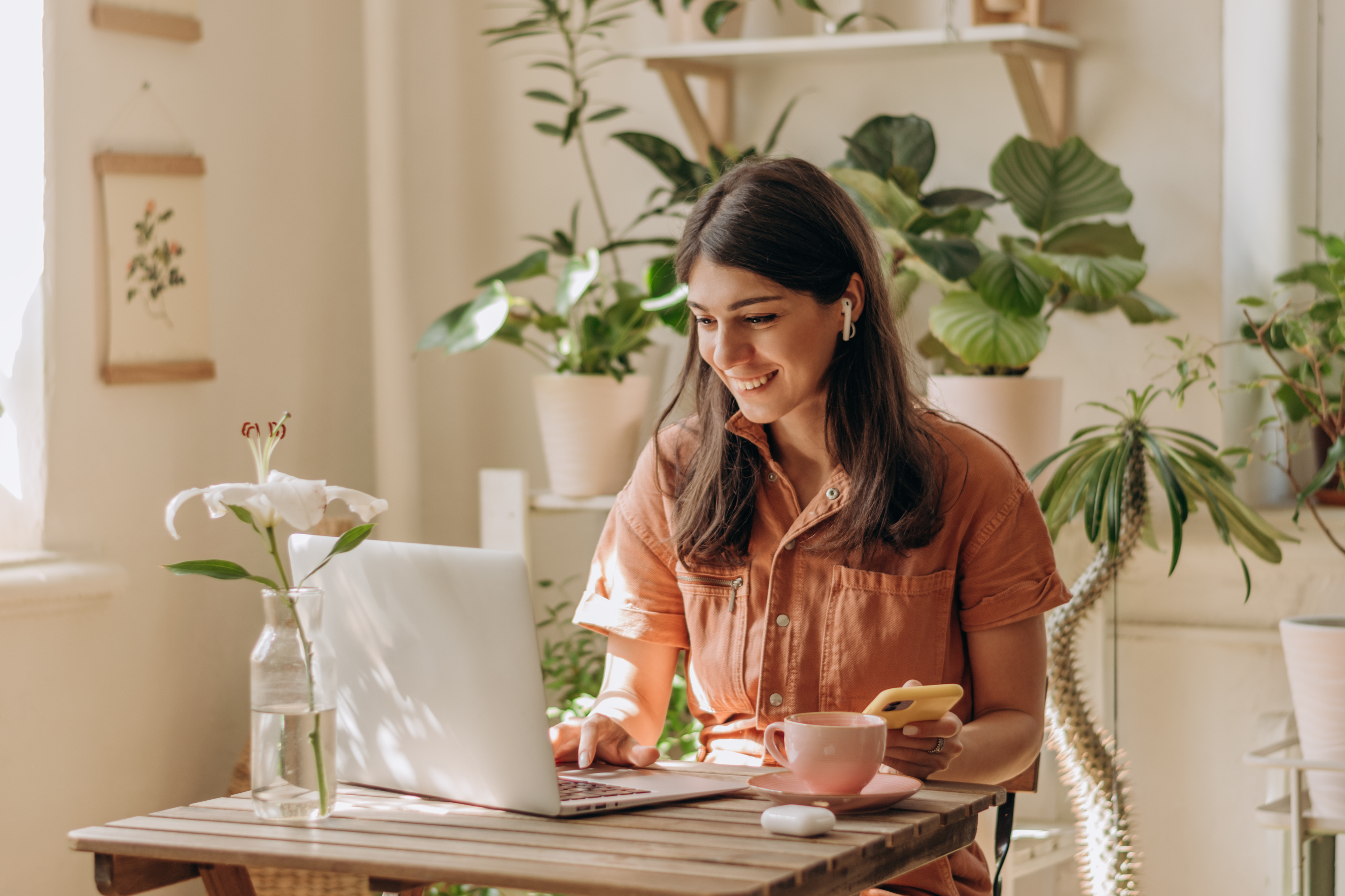 Woman sits at a table and uses a laptop. Behind her are shelves of houseplants.