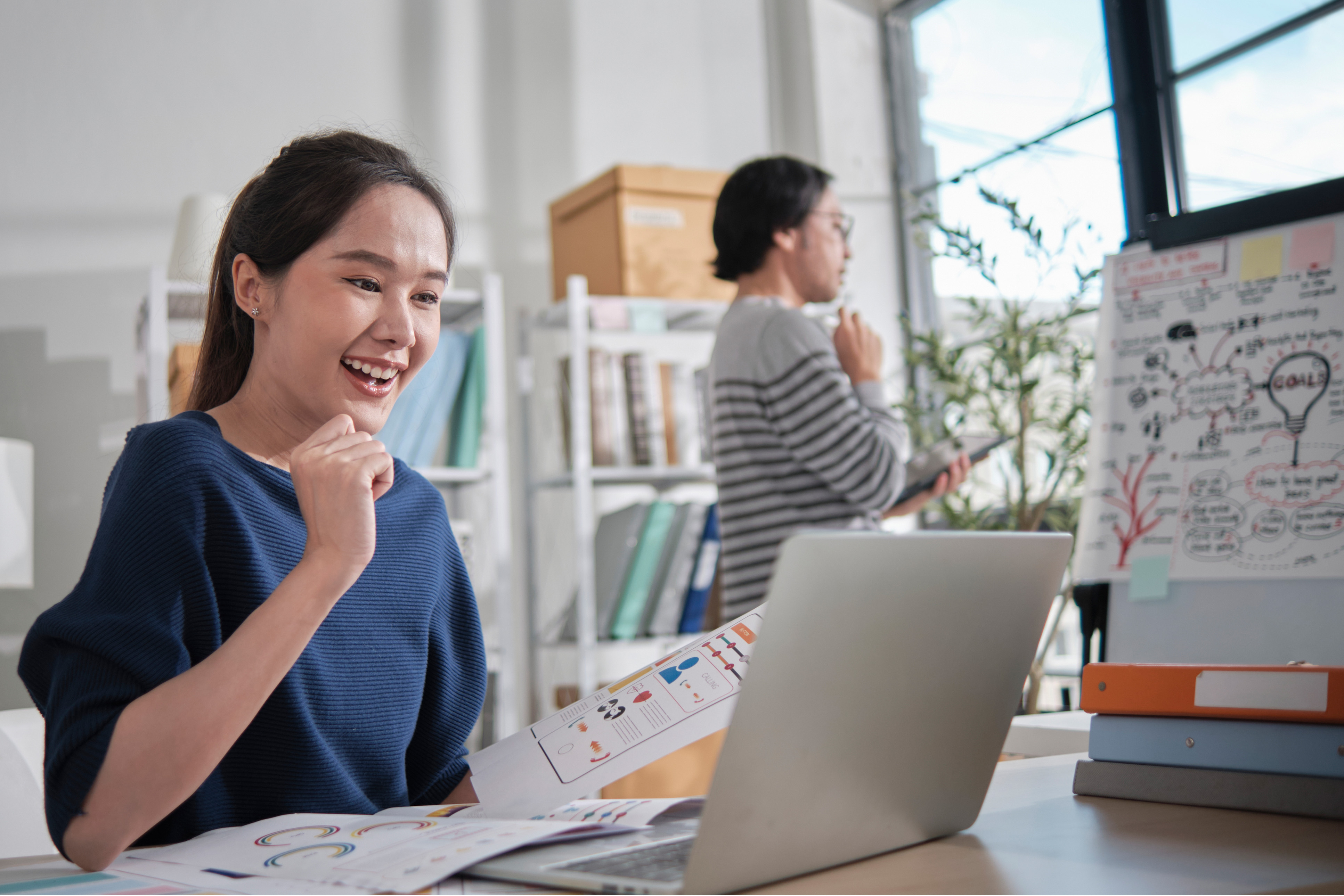 Woman uses a laptop, man behind her stares out of a window pondering the scene below.